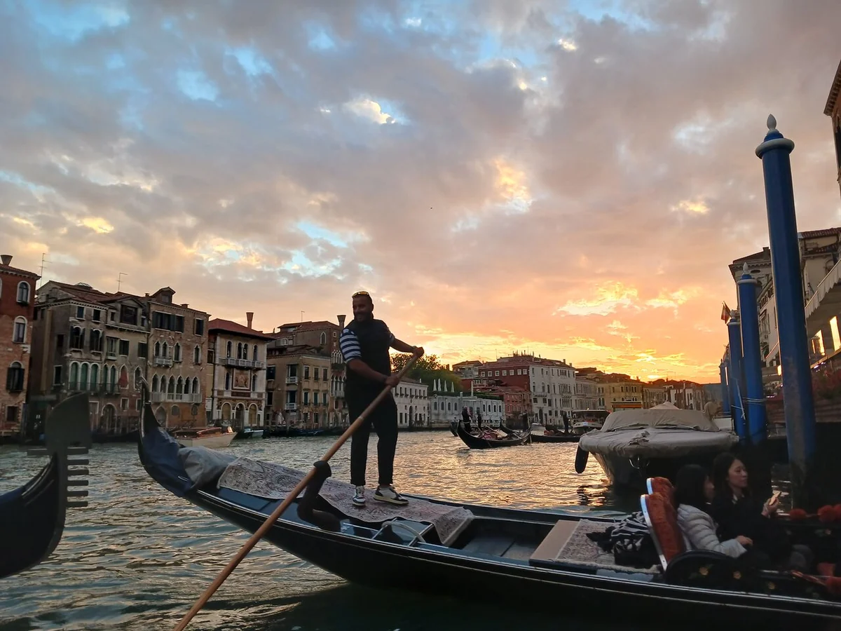A gondolier rowing a gondola past palazzi under an orange sunset sky